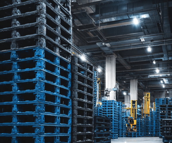 The interior of a large, industrial warehouse featuring tall stacks of empty blue and black reusable plastic pallets, with a yellow forklift visible in the background.